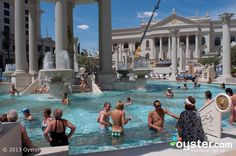 many people are in the water at an outdoor swimming pool with fountains and columns on either side