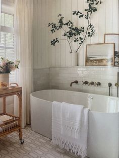 a white bath tub sitting under a window next to a wooden table with flowers on it