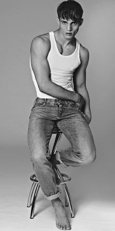 a young man sitting on top of a stool wearing jeans and a tank top with his hands in his pockets