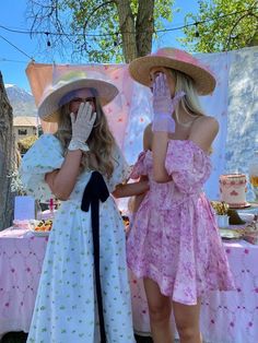 two women in dresses and hats standing next to each other near a table with food on it