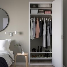 a white closet with clothes and shoes hanging on the wall next to a bed in front of a round mirror