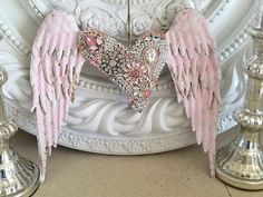 an angel heart with pink wings on a white pedestal next to two silver candlesticks