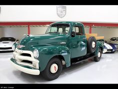 an old green truck parked in a garage next to other classic cars on the floor