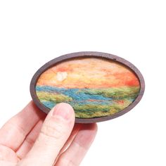 a hand holding up a small wooden brooch with a painting on it's side