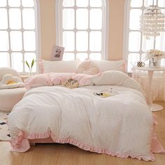 a white bed with pink ruffles and pillows in a room next to two windows