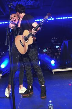 two people standing next to each other while one person holds a guitar and sings into the microphone