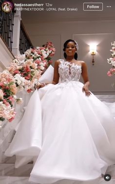 a woman in a white wedding dress is walking down the stairs with flowers behind her