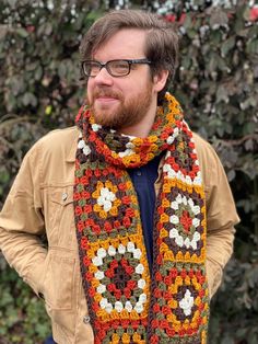 a man wearing glasses and a crocheted scarf stands in front of some bushes