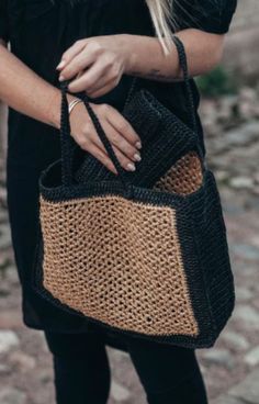 a woman holding a black and tan woven tote bag on her left hand while standing in the street