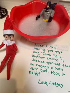 an elf is sitting in front of a red bowl filled with water and two toy batman figures