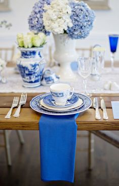 a blue and white table setting with flowers in vases on the side, silverware at the end