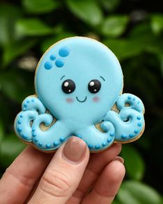 a hand holding a cookie shaped like an octopus with blue icing on it's face