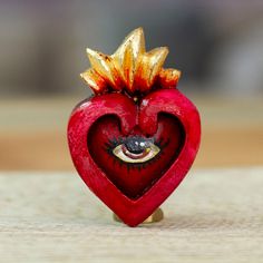 a red heart shaped object with an eye on it's side and gold leaves sticking out of the top