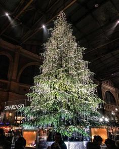 a large christmas tree is lit up in the middle of a building with people standing around it