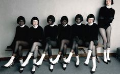 a group of women sitting next to each other on top of a wooden bench in front of a wall