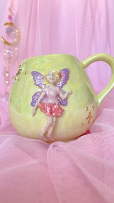 a green mug with a little fairy on it's side sitting on a pink surface