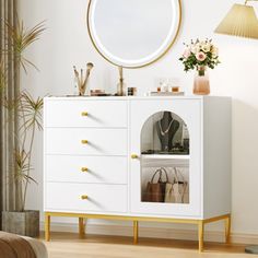 a white dresser with gold handles and drawers in a room next to a lamp, mirror and potted plant