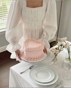 a woman is holding a pink heart shaped cake on a white table with flowers in vases