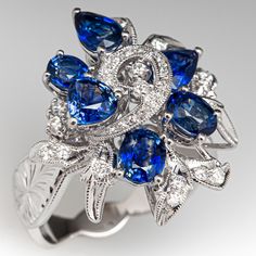 This impressive cluster style ring is accented with three (3), prong set, oval mixed cut natural sapphires, two (2), prong set, pear mixed cut natural sapphires and one (1), prong set, triangular mixed cut natural sapphire. The sapphires are bordered with four (4), prong set, round brilliant cut diamonds and twenty- eight (28), bead set, round brilliant/single cut diamonds, bordered with milgrain edging. Engraved details accent the shoulders of the shank. The ring measures 22.5mm at the top, rises 10.1mm above the finger, tapering to 3.6mm wide and 1.2mm thick at the base of the shank. This ring is currently a size 6.75. Luxury Sapphire Cubic Zirconia Cluster Ring, Luxury Sapphire Open Ring Jewelry, Blue Sapphire Diamond, Bead Set, One 1, Natural Sapphire, Diamond Cluster, Sapphire Diamond, Round Brilliant Cut Diamond