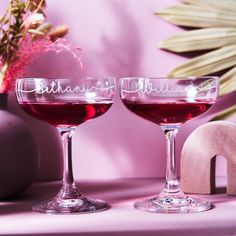 two glasses of red wine sitting next to each other on a pink tablecloth with flowers in the background