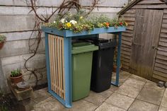 two trash cans sitting next to each other in front of a wooden shed with plants growing out of them