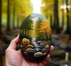 a hand holding an easter egg painted with trees and rocks in the forest, on a sunny day