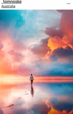 a man walking on the beach in front of an orange and blue sky with clouds