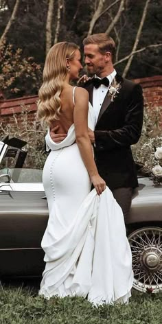 a bride and groom standing in front of an old car with the bride's dress flowing down