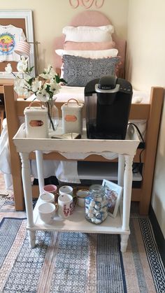 a small white table with cups and mugs on it in front of a bed