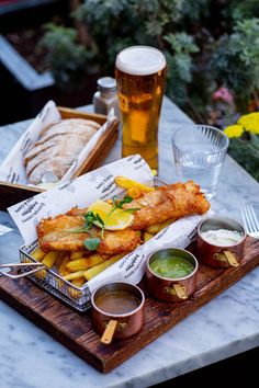 fish and chips on a wooden platter next to a glass of beer, bread and dips