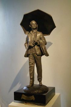 a bronze statue of a man holding an umbrella on top of a white pedestal in front of a wall