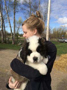 a woman holding a black and white puppy in her arms while standing on a dirt road