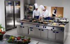 two chefs preparing food in a kitchen with stainless steel cabinets and counters, surrounded by fresh vegetables