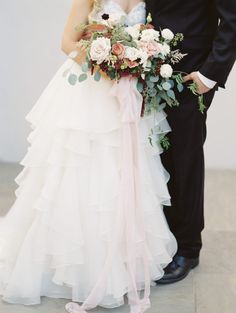 a bride and groom pose for a wedding photo in front of a white wall with flowers