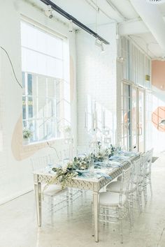 a long table with white chairs and flowers on it in front of a large window