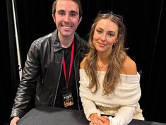 a man and woman sitting at a table smiling for the camera with black curtains behind them