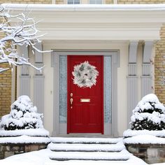 a red door with a wreath on it in front of snow covered bushes and trees