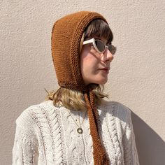 a woman wearing sunglasses and a knitted hat with a scarf around her neck is standing in front of a white wall