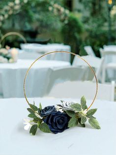 a blue rose and greenery in a gold hoop on a table with white linens