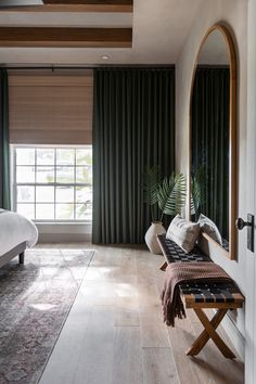 a bedroom with a bed, mirror and bench in front of a window that has green curtains