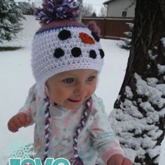 a baby wearing a crocheted hat standing next to a tree in the snow