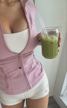 a woman in white shorts and pink jacket holding a glass with green smoothie inside