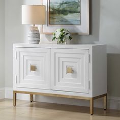 a white sideboard with two doors and a vase on top, in front of a painting