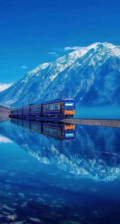 a blue and yellow train traveling down tracks next to a large body of water with snow covered mountains in the background