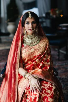 a woman in a red and gold bridal outfit sitting on a chair with her hands clasped to her chest