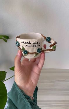 a hand holding a ceramic cup with writing on the bottom and green leaves around it