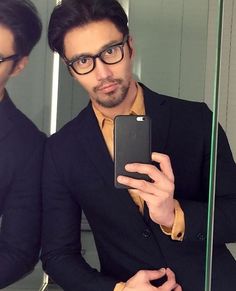 a man wearing glasses is looking at his cell phone in front of a bathroom mirror