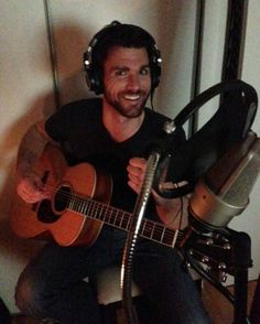 a man sitting in front of a microphone and holding an acoustic guitar with headphones on