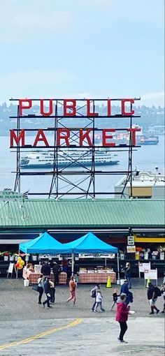 people are walking around in front of the public market on a sunny day with boats in the background