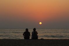 two people sitting on the beach watching the sun set